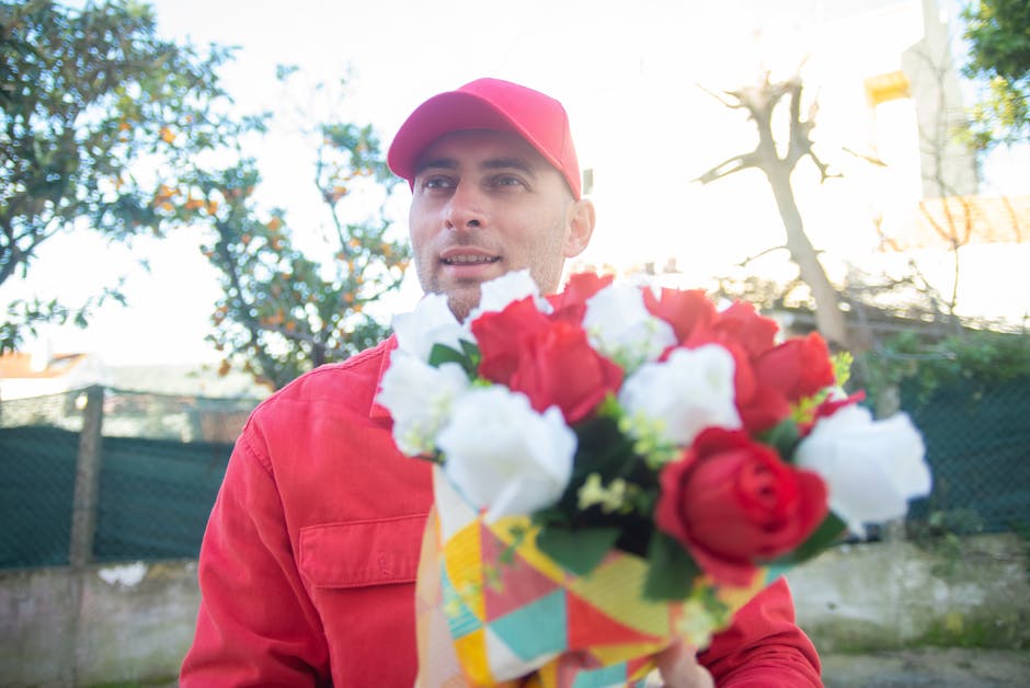 A man dressed in a red jacket and matching red cap holds a vibrant bouquet of fresh flowers, including red and white blooms, wrapped in colorful patterned paper. He is outdoors in natural daylight, wi