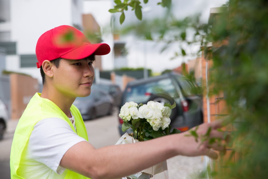 A young male flower delivery person wearing a red cap and high-visibility yellow vest is placing a bouquet of white hydrangeas wrapped in white paper next to a green leafy shrub on an outdoor resident