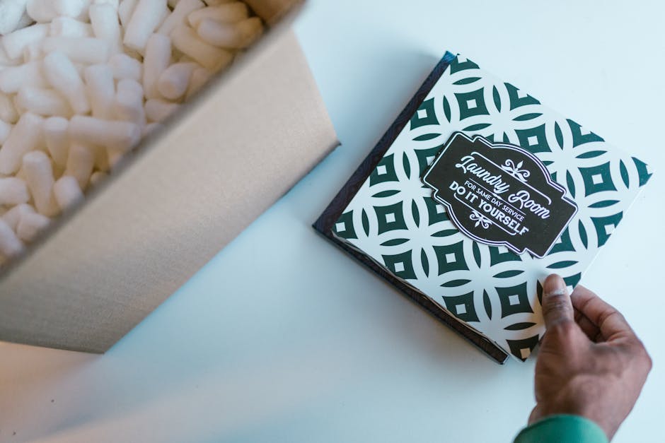 A close-up of a person’s hand holding a black and white patterned box with the label 'Laundry Room for same day service, do it yourself,' positioned on a white surface next to a partially visible be
