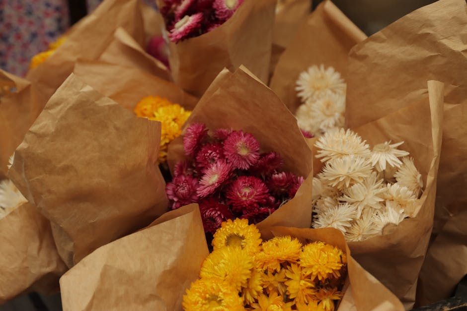 A selection of freshly cut flowers arranged in brown paper wraps, featuring vibrant pink chrysanthemums with fluffy petals, bright yellow daisies with numerous small petals, and elegant white blooms r