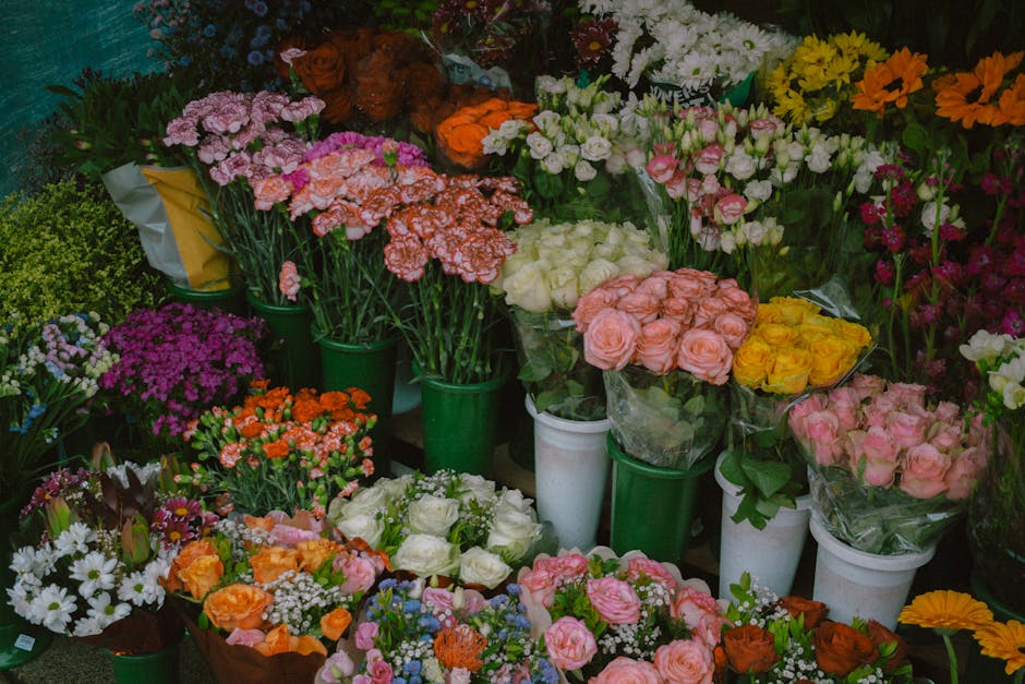 A vibrant display of fresh flowers arranged in various containers, including white and green vases and buckets. The collection features roses in shades of pink, peach, and yellow, grouped together in 